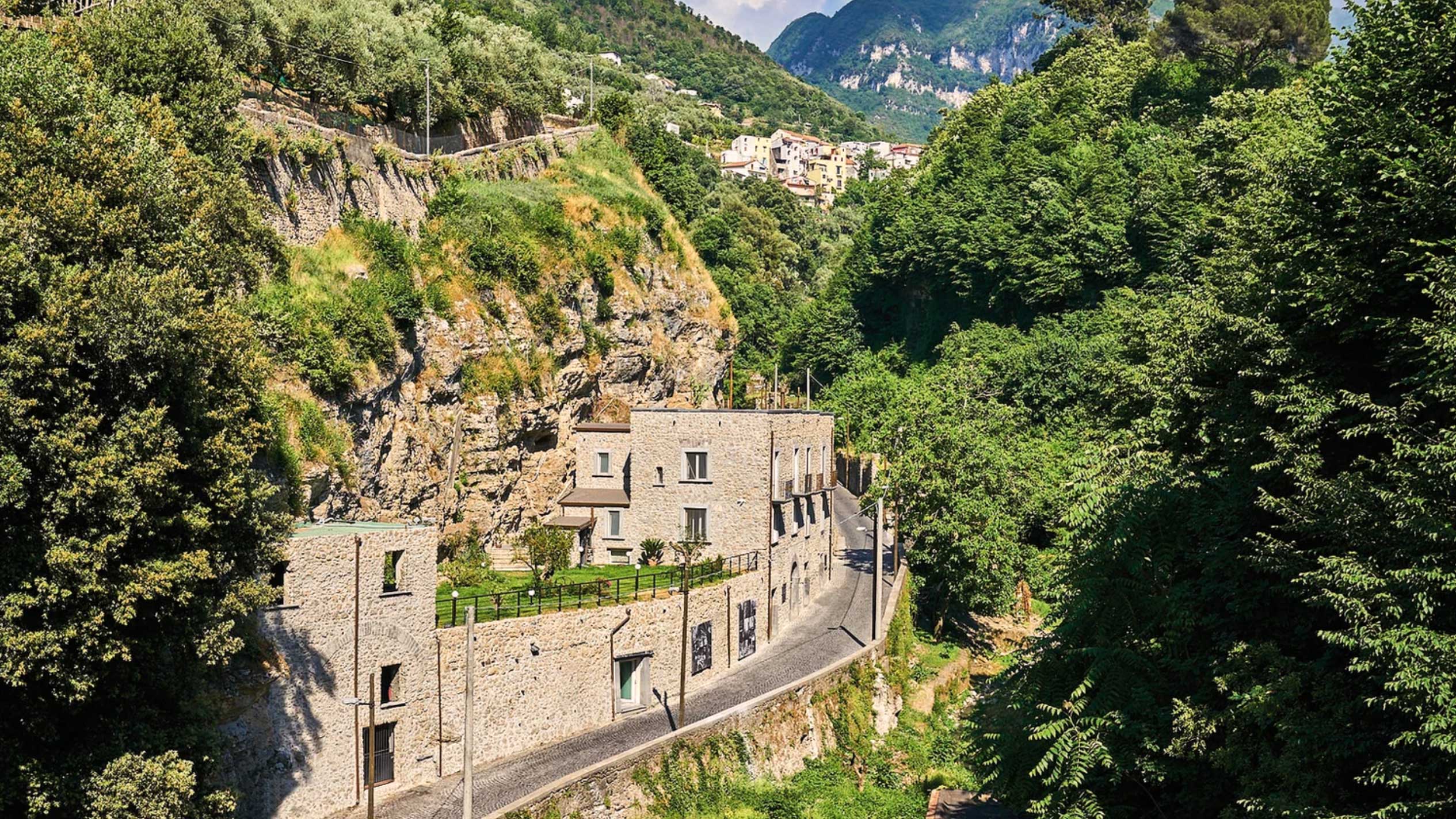Historisches Gebäude von Pastificio Gentile in einer üppig bewachsenen Tal-Landschaft, das traditionelle Handwerkskunst und regionale Identität verkörpert. Die Architektur fügt sich harmonisch in die Natur ein und repräsentiert die Ursprünge des Unternehmens in Gragnano