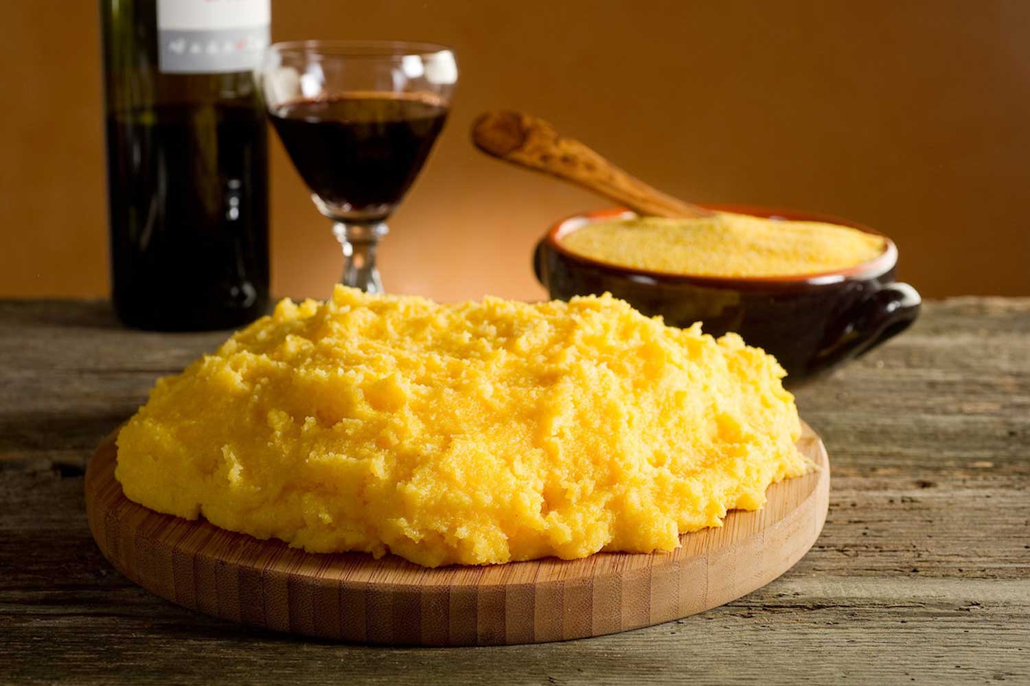 Ein rustikales Arrangement mit frisch zubereiteter Polenta, angerichtet auf einem runden Holzbrett. Im Hintergrund steht eine traditionelle schwarze Schale mit Polenta und einem Holzlöffel, sowie ein Glas tiefroten Weins und eine Weinflasche, die das italienische Flair unterstreichen. Die Szene ist auf einem rustikalen Holztisch präsentiert, mit warmem Licht, das die leuchtend gelbe Farbe der Polenta hervorhebt. Dieses Bild vermittelt die Authentizität und den Genuss italienischer Tradition.