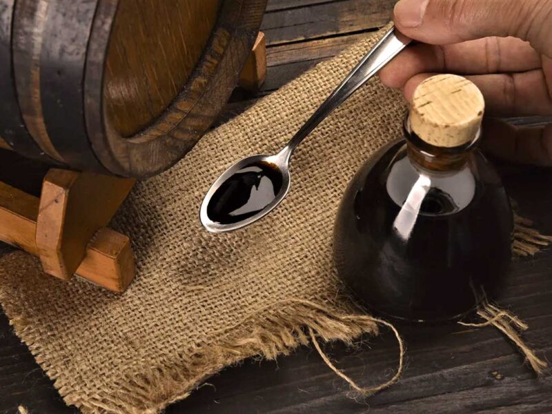 Ein Löffel mit dickflüssigem, dunklem Aceto Balsamico vor einem traditionellen Holzfass und einer kleinen Glasflasche mit Korkverschluss. Der gereifte Essig ruht auf einem rustikalen Jutesack, was die handwerkliche Tradition und Qualität des italienischen Balsamico-Essigs unterstreicht.