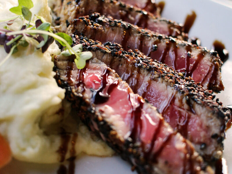 Ein Teller mit in Sesamkruste gebratenem, rosa-gegartem Thunfischfilet, serviert mit Kartoffelpüree und mit Aceto Balsamico beträufelt.