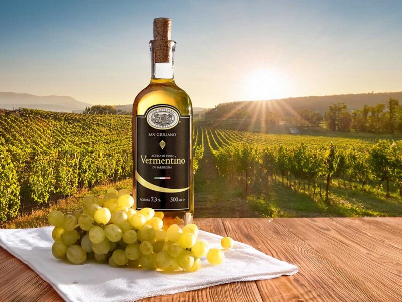 Eine Flasche “Aceto di Vino Vermentino” vor einer malerischen Weinlandschaft mit grünem Rebstock und untergehender Sonne.