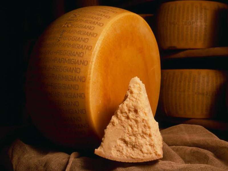 Ein großes, traditionell geprägtes Laib Parmigiano Reggiano mit einem herausgebrochenen Stück, das die körnige, reife Textur dieses berühmten italienischen Hartkäses zeigt. Im Hintergrund sind weitere gereifte Laibe in einem Käselager zu sehen. Diese Aufnahme dient als Titelbild für einen neuen Artikel in der Centro Italia Gazzetta über die Herkunft, Herstellung und kulinarische Bedeutung von Parmigiano Reggiano.