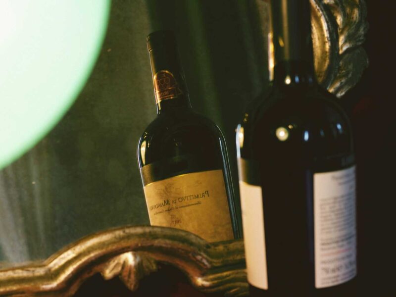 Stimmungsvolle Aufnahme einer Flasche Primitivo di Manduria, die sich in einem antiken Spiegel spiegelt. Der apulische Rotwein, bekannt für seine intensiven Aromen und kräftigen Noten, steht im Fokus eines neuen Artikels der Centro Italia Gazzetta über die Faszination und Geschichte des Primitivo-Weins.