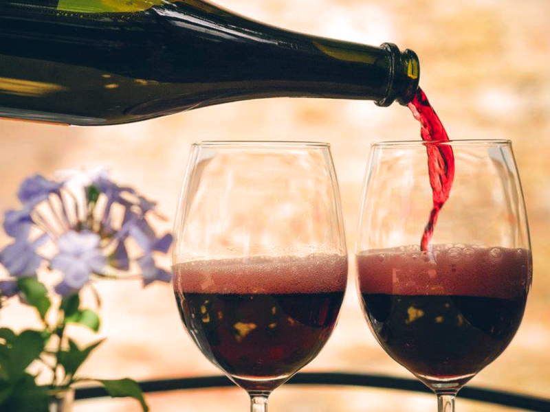 Eine Flasche Lambrusco, aus der perlender roter Schaumwein in zwei Weingläser gegossen wird. Im Hintergrund sanfte, mediterrane Farben und violette Blüten – eine Hommage an den ikonischen italienischen Lambrusco.