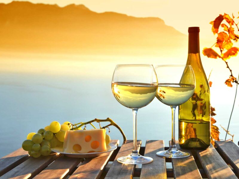 Zwei Gläser Lugana-Weißwein stehen auf einem Holztisch mit einer Flasche Wein, grünen Trauben und einem Stück Käse, während die Abendsonne den Gardasee und die umliegenden Berge in ein goldenes Licht taucht.