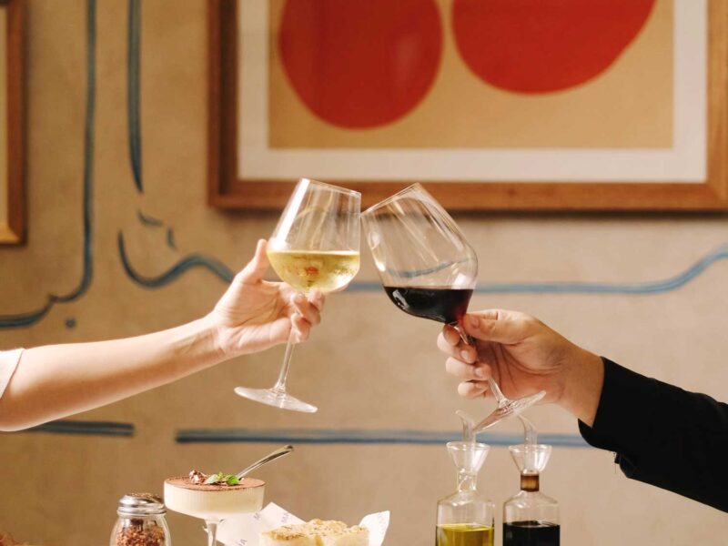 Ein stilvoll gedeckter Tisch in einem italienischen Restaurant, auf dem eine klassische Pizza mit Basilikum serviert wird. Zwei Personen stoßen mit einem Glas Rot- und Weißwein an – ein eleganter Moment mediterraner Lebensfreude.