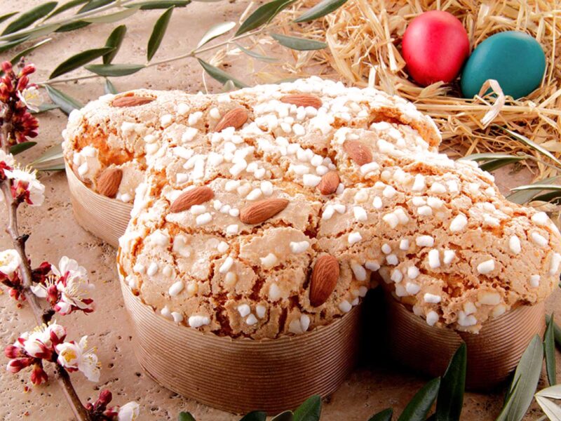 Traditionelle italienische Colomba Pasquale, ein Ostergebäck in Taubenform, mit knuspriger Mandel- und Zuckerglasur. Im Hintergrund dekorative Olivenzweige und bunte Ostereier auf Stroh