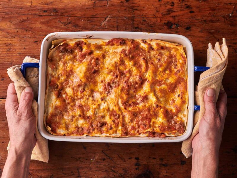 Ein frisch gebackener Lasagne-Auflauf in einer weißen Keramikform auf einem rustikalen Holztisch, gehalten von zwei Händen mit Topflappen.