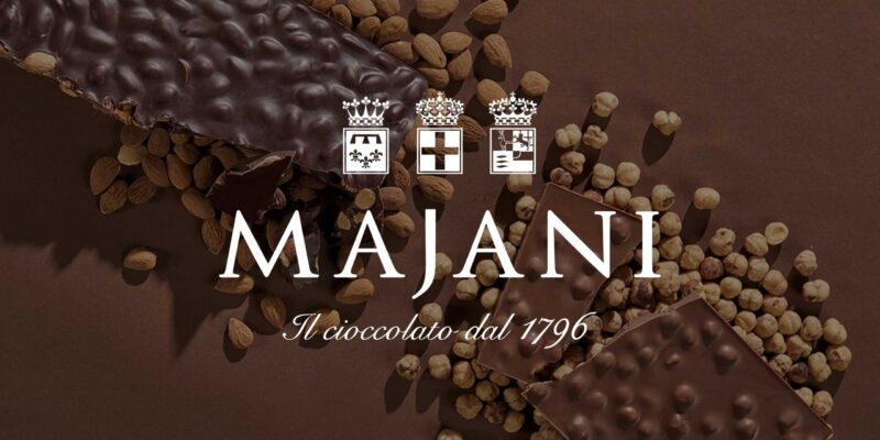 Majani-Schokoladentafeln mit ganzen Haselnüssen und Mandeln auf braunem Hintergrund, begleitet vom Majani-Logo und dem Schriftzug „Il cioccolato dal 1796“ – italienischer Traditionshersteller feinster Schokolade seit über 200 Jahren.