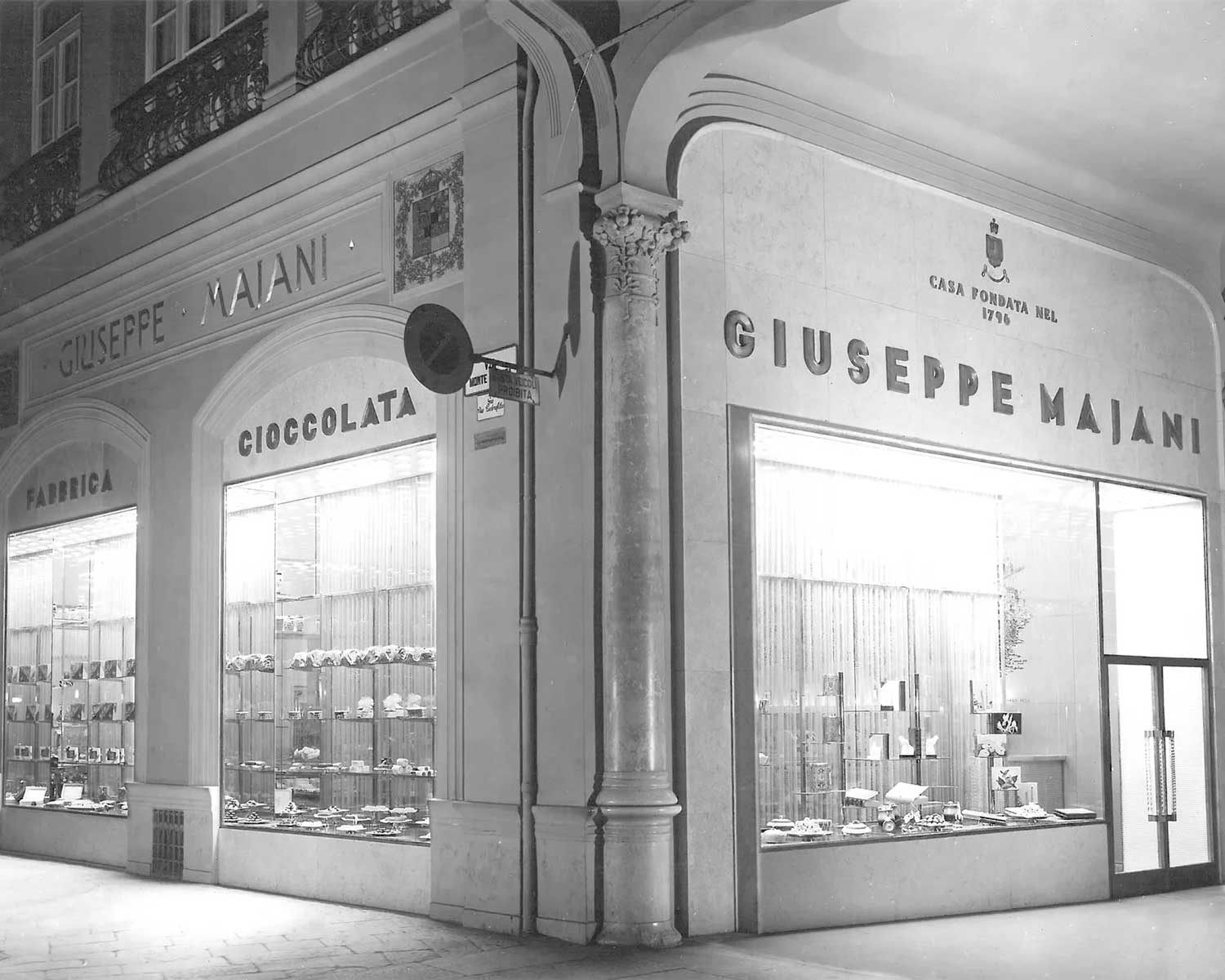 Schwarzweißfoto des historischen Schokoladengeschäfts von Giuseppe Majani in Bologna mit beleuchtetem Schaufenster und klassischer Architektur.