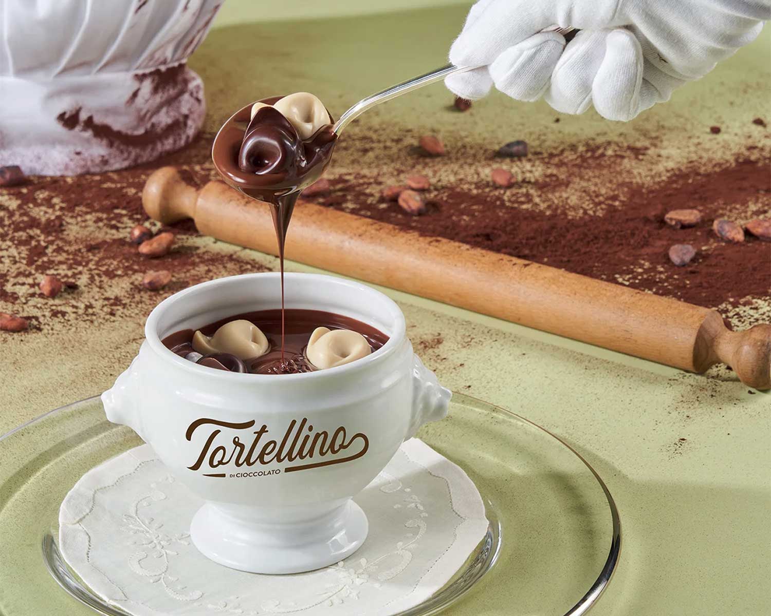 Nahaufnahme eines weißen Schälchens mit der Aufschrift “Tortellino di Cioccolato”, gefüllt mit flüssiger Schokolade und kleinen Schokoladentortellini, auf einem mit Kakaopulver bestäubten Tisch.