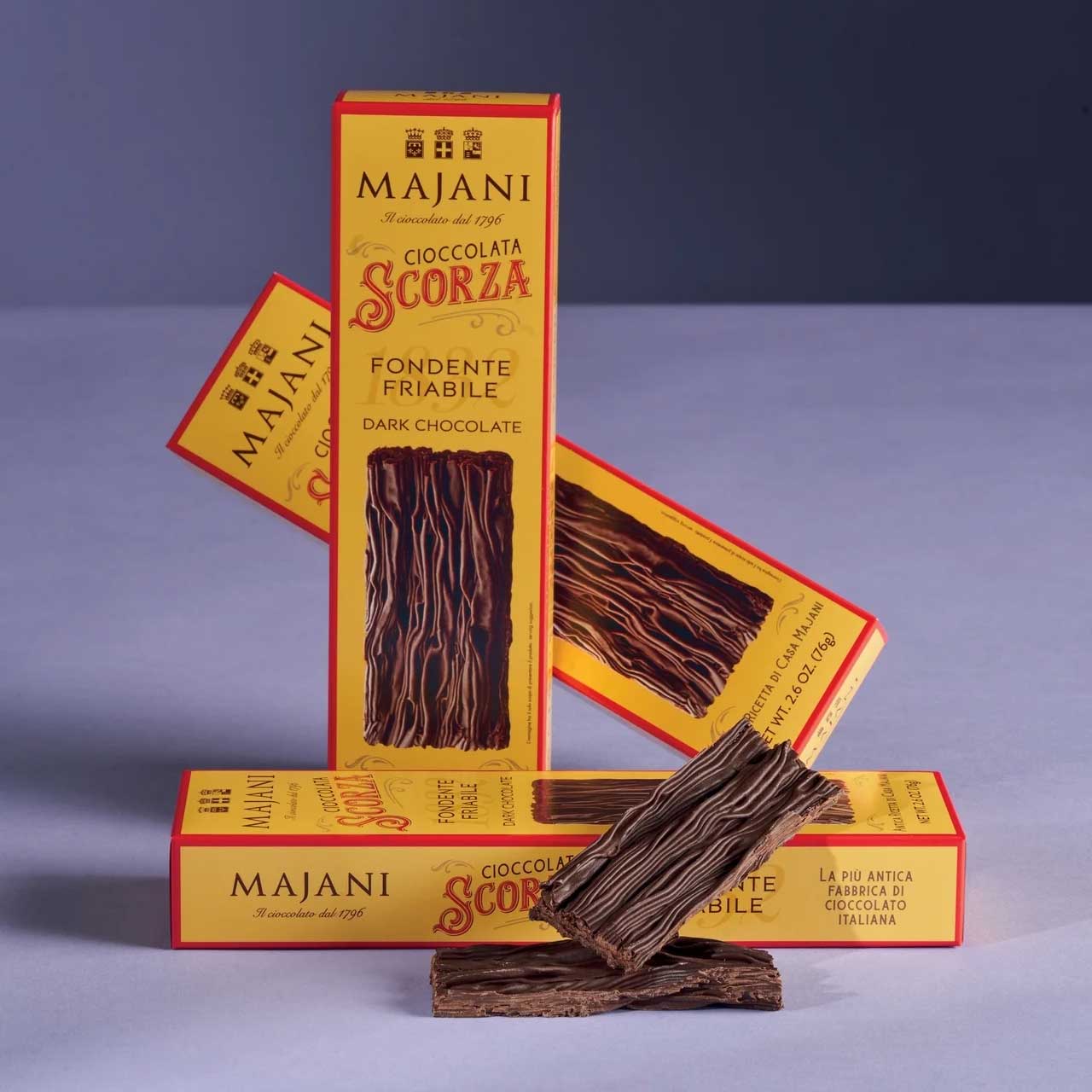 Verpackung der traditionellen „Cioccolata Scorza“ von Majani, eine dunkle, brüchige Schokolade mit charakteristischer Rindenstruktur, aufgestellt auf einem hellblauen Hintergrund mit geöffneten Tafeln im Vordergrund.