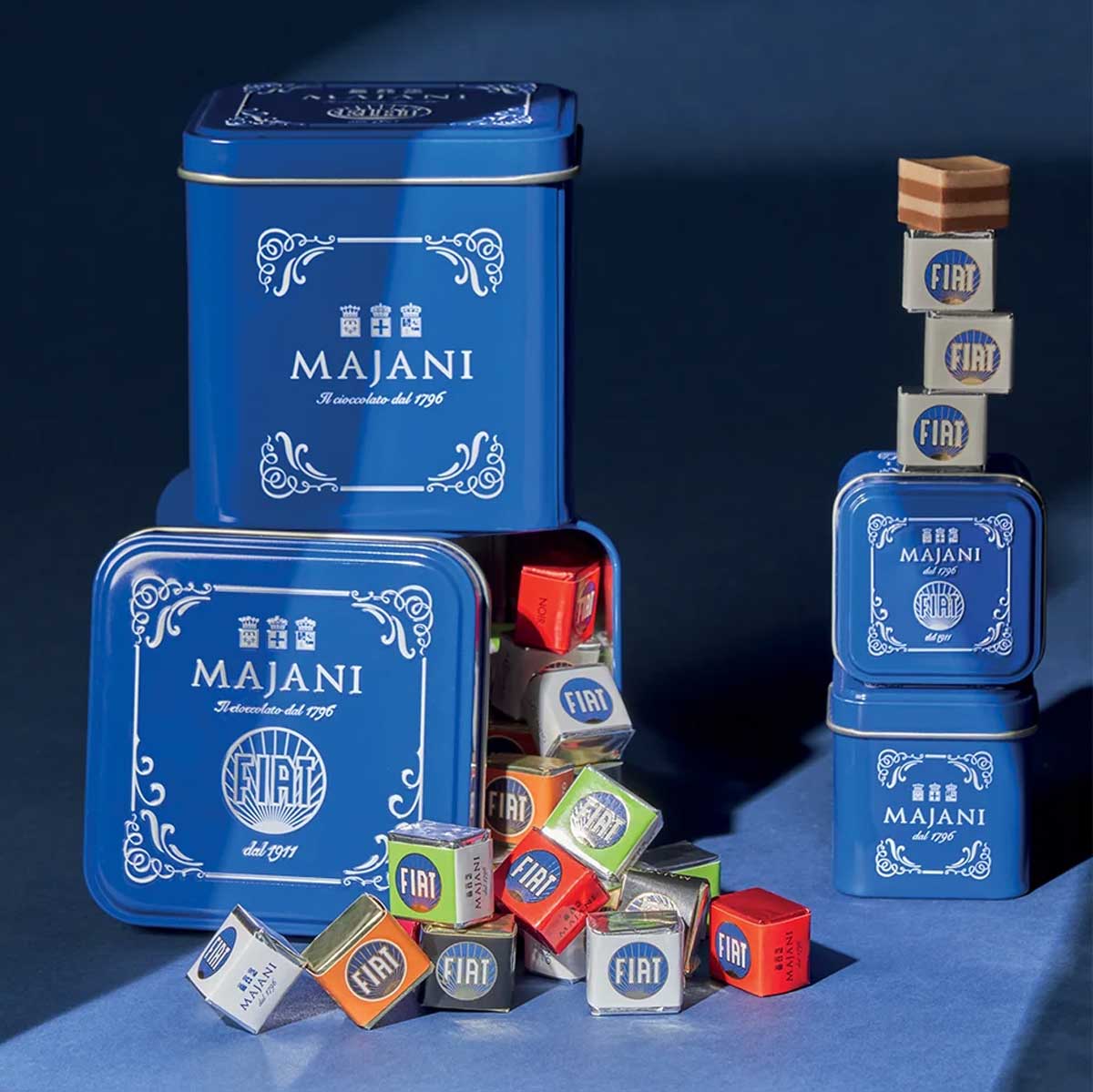 Elegante blaue Blechdosen von Majani mit „Fiat“-Logo, gefüllt mit bunten Schokoladenwürfeln, stilvoll arrangiert auf dunkelblauem Hintergrund – eine Hommage an das historische Fiat-Schokoladenpralinen-Rezept von 1911.