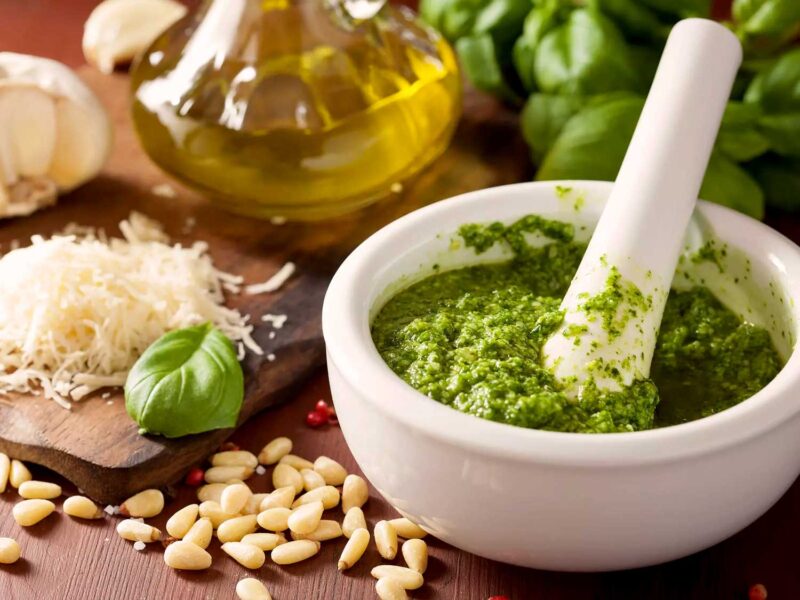 Traditionelle Zutaten für Pesto alla Genovese: Frischer Basilikum, geriebener Käse, Knoblauch, Pinienkerne und Olivenöl neben einem weißen Mörser mit grünem Pesto.