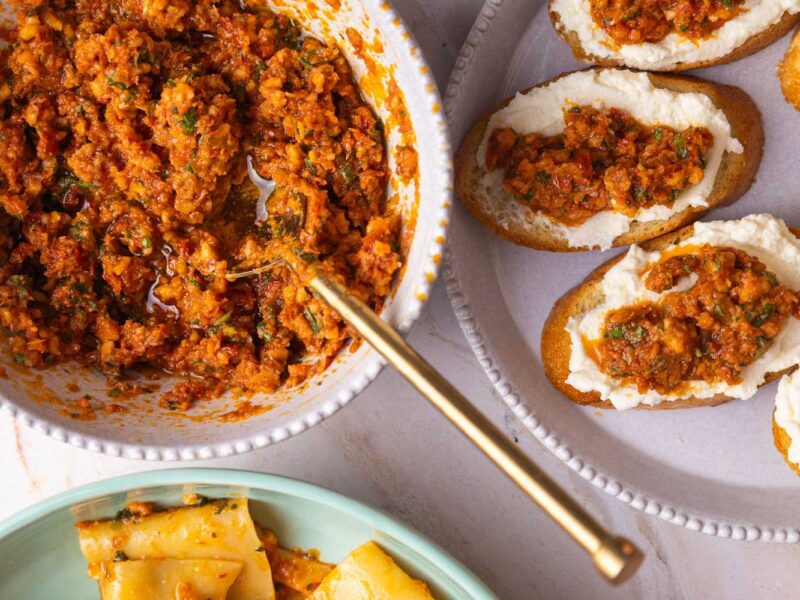 Pesto Rosso auf knusprigen Crostini mit Ricotta, daneben eine Schale mit der frischen Paste und ein Teller mit Pasta – rustikale mediterrane Präsentation.