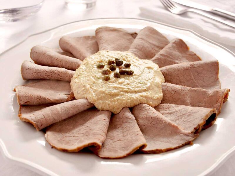Elegante Präsentation von Vitello Tonnato mit dünnen Kalbfleischscheiben, die fächerförmig um eine Thunfisch-Kapern-Sauce arrangiert sind, serviert auf einem weißen Porzellanteller.
