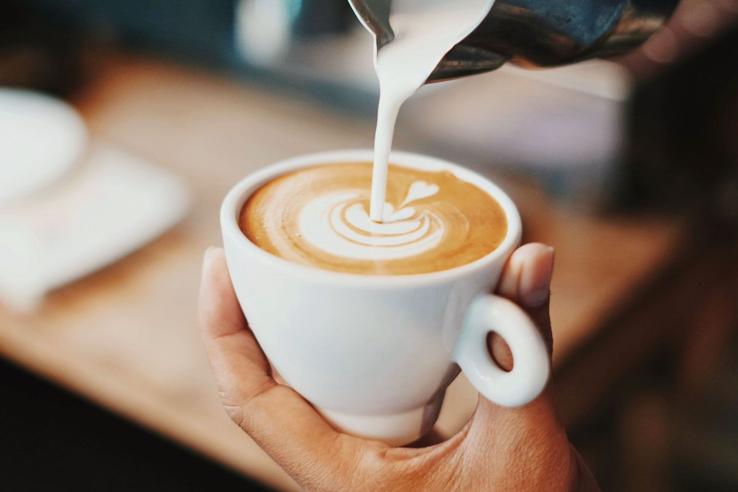 Detailaufnahme eines Cappuccinos mit Latte Art, während frische Milch aus einem Kännchen in die Tasse gegossen wird – ein Moment der Barista-Kunst.