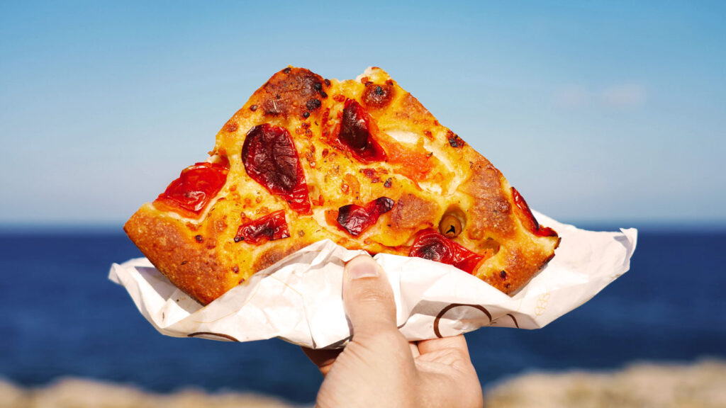 Ein Stück Focaccia mit Paprika wird vor dem Meer in die Luft gehalten – Streetfood-Genuss am Wasser