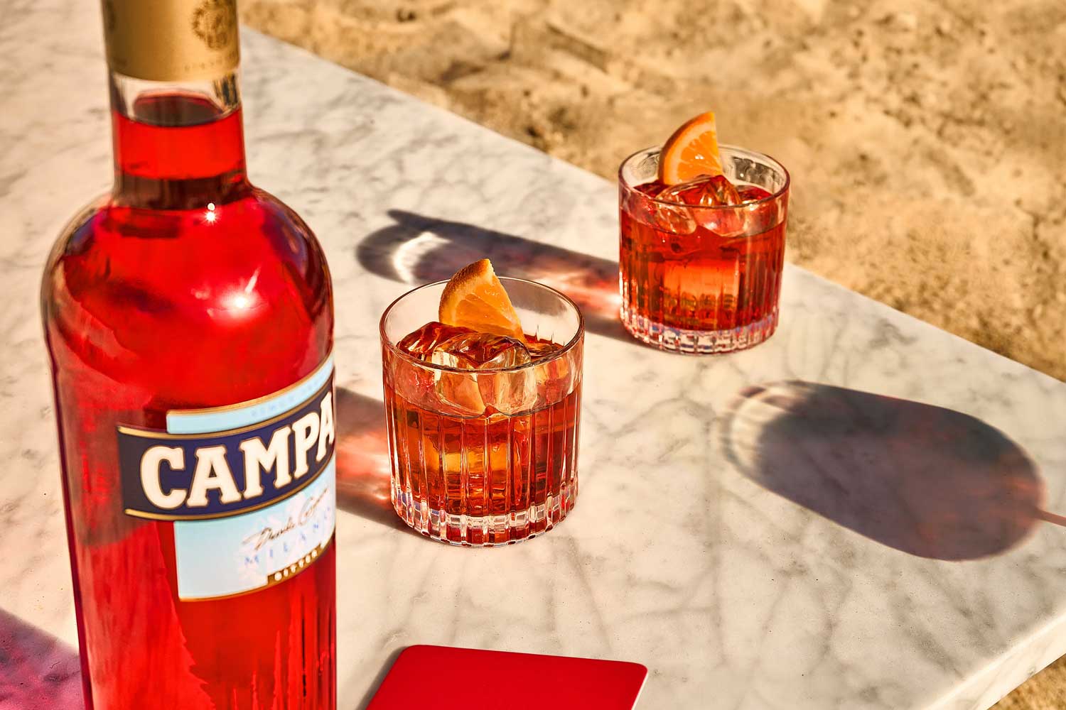Negroni & Negroni Sbagliato - Der bittersüße Kult-Cocktail aus Italien Zwei Gläser Negroni mit Eiswürfeln und Orangenscheiben auf einem Marmortisch am Strand, neben einer Flasche Campari.