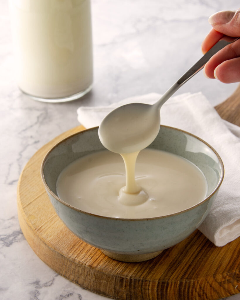 Löffel, der eine seidige Besciamella-Sauce aus einer Keramikschale schöpft, mit Milchflasche im Hintergrund