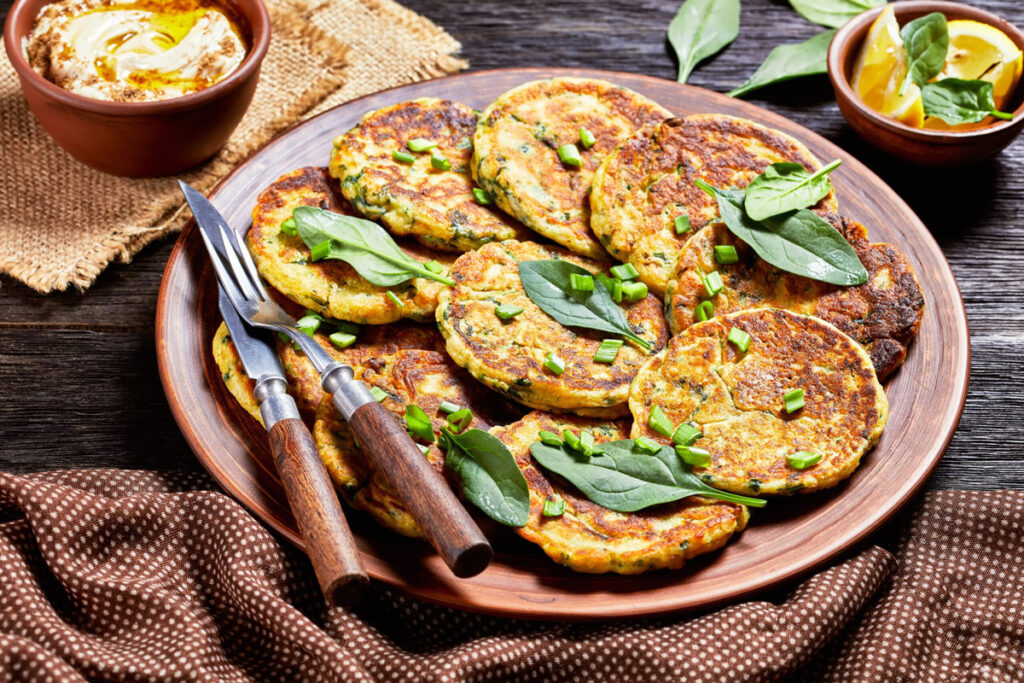 Frittelle aus Kichererbsenmehl angerichtet auf einem rustikalen Holzteller mit frischen Kräutern, serviert mit cremigem Hummus und Zitronenspalten.