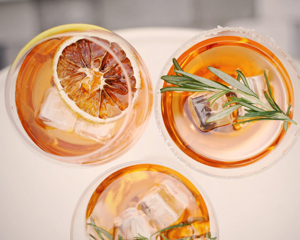 Draufsicht auf drei elegante Gläser mit Spritz, verziert mit Eiswürfeln, getrockneten Orangenscheiben und frischem Rosmarin – ein moderner Twist des Klassikers.