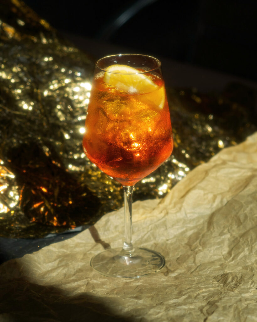 Ein leuchtend orangefarbener Spritz mit Zitronenscheibe im eleganten Glas, im dramatischen Licht vor einer goldenen, festlichen Kulisse.